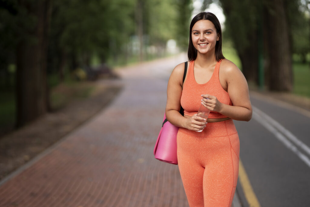 "Smiling woman in an orange workout outfit walking in a park with a yoga mat and water bottle, representing a renewed sense of movement and vitality after 50."